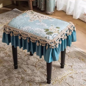 May include: A small, upholstered stool with a light blue seat featuring floral embroidery and a scalloped lace trim. The stool has a matching blue skirt and dark brown legs, suitable for a bedroom or vanity.