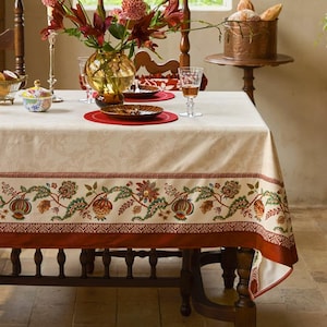 Op de afbeelding: Een crèmekleurig tafelkleed met een gedetailleerde bloemenrand in rood, groen en bruin. De tafel is gedekt met borden, glazen en een vaas met bloemen, wat een elegante eettafereel creëert. De tafel is van donker hout.