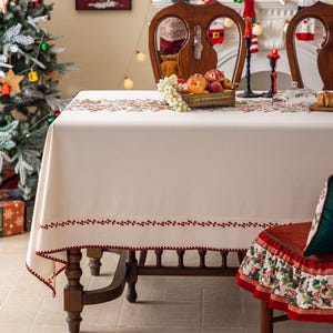 Puede incluir: Un mantel color crema con un borde bordado en rojo, preparado para una ocasión festiva. La mesa está adornada con una cesta de frutas, velas y un árbol de Navidad al fondo. La mesa es de madera.