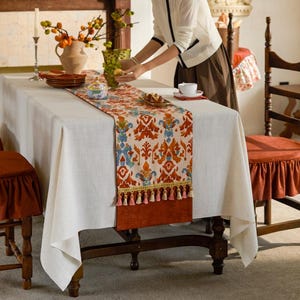May include: A dining table set for a meal. The table is covered with a white tablecloth and a patterned table runner with orange, blue, and cream colors. The runner has tassels at the end. There are plates, a vase, and a person setting the table.