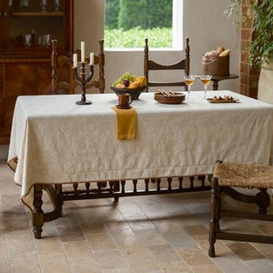 以下が含まれることがあります： 食事のためにセットされたダイニングテーブル。テーブルは、装飾的なボーダーが付いたクリーム色のテーブルクロスで覆われています。燭台、フルーツボウル、プレート、グラスがテーブルに配置されています。木製の椅子とスツールが近くにあります。