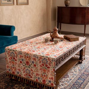 May include: A rectangular coffee table with a floral patterned tablecloth. The tablecloth has a cream background with red, blue, and orange flowers. The table has a dark wood finish and is decorated with a copper teapot, cups, and a book. The tablecloth has tassels.