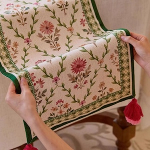 May include: Cream-coloured floral table runner with a pattern of pink and red flowers, green leaves, and a green border. The runner has red tassels at the corners. A decorative piece for a dining table.