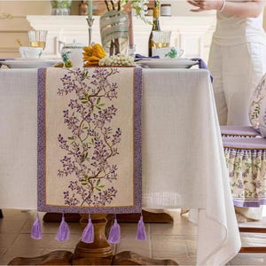 May include: A white linen tablecloth with a purple floral patterned table runner. The table runner has tassels on each end and features a delicate floral design with purple flowers and green leaves.