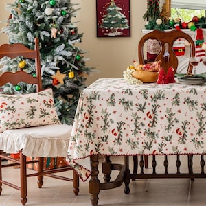 Op de afbeelding: Een feestelijke kerstscène met een versierde boom, een houten stoel met een patroon kussen en een tafel gedekt met een feestelijk tafelkleed. Het tafelkleed heeft een herhalend patroon van kerstbomen, zuurstokken en kerstmutsen.