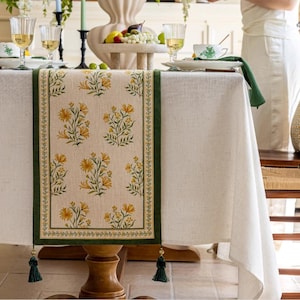 Könnte beinhalten: Eine weiße Tischdecke mit einem grünen und gelben Tischläufer mit Blumenmuster. Der Tischläufer hat an jedem Ende Quasten.
