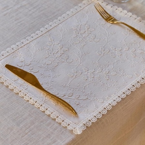 May include: A white, square placemat with an embroidered floral design and a delicate lace trim. A gold-colored fork and knife are placed on the placemat, creating an elegant table setting. The placemat is on a beige tablecloth.