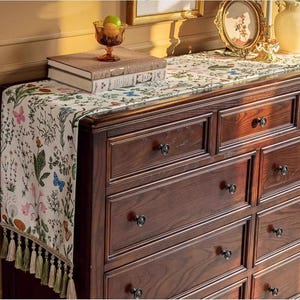 May include: A white chest of drawers with six drawers and a dark brown wood finish. The chest of drawers has black metal knobs. A white table runner with a floral pattern and green tassels is on top of the chest of drawers.