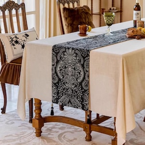 May include: A dining table set with a cream-colored tablecloth and a black and white patterned table runner. The runner features a detailed floral design. A decorative pillow with a floral design rests on a wooden chair.