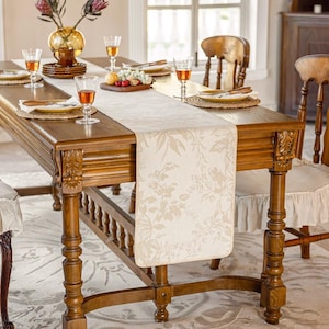 May include: A wooden dining table set for a meal, with a cream floral table runner. The table is set with plates, glasses, and a vase of flowers. The chairs have decorative backs and ruffled seat covers.