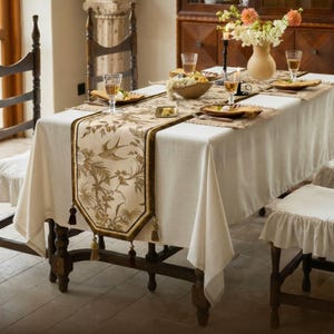 May include: A rectangular dining table covered with a plain white tablecloth and a long, beige table runner featuring a brown botanical print with birds and flowers. The runner is finished with decorative tassels at both ends and edges.