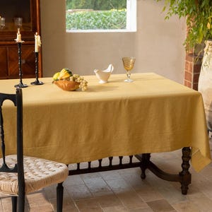 May include: A mustard-yellow rectangular tablecloth covers a dark wooden table. The table is set with a decorative bowl of fruit, a white gravy boat, and a textured glass. Two black candle holders with lit candles are on the left.