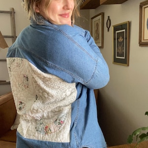 May include: A long-sleeved blue denim shirt featuring a large, rectangular back panel made of white quilted fabric with a floral pattern and textured lace detailing. The shirt has a classic collar, button-down front, and a curved hemline.