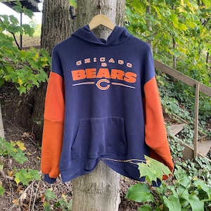 May include: A navy blue hooded sweatshirt with bright orange sleeves and a front kangaroo pocket. The chest features the text CHICAGO BEARS in orange and white lettering, along with a small NFL logo and the orange Chicago Bears C logo centered below.