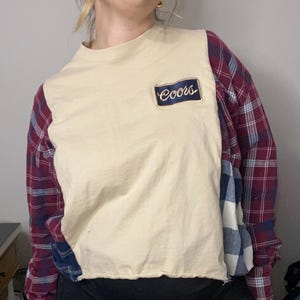 Könnte beinhalten: Ein cremefarbenes, kurzärmeliges Shirt mit einem Coors-Aufnäher auf der Vorderseite. Die Ärmel sind aus rot-blau kariertem Flanellstoff gefertigt. Das Shirt hat einen rohen, unfertigen Saum.