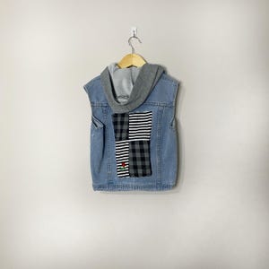 May include: A light blue denim waistcoat featuring a grey jersey hood and a central back panel composed of four alternating squares of black and white plaid and striped fabric, with a small embroidered red rose detail on the lower left square.