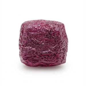 May include: A raw, deep magenta-colored gemstone with a rough, textured surface and an irregular, blocky shape. The stone exhibits a translucent quality with varying depths of red and purple tones across its multifaceted, uneven exterior.
