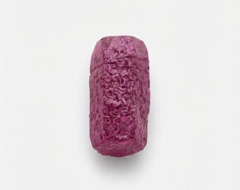 May include: A raw, unpolished ruby crystal with a deep magenta-pink hue. The gemstone features a hexagonal, elongated columnar shape with a rough, textured surface showing natural crystalline facets and irregular edges throughout its structure.