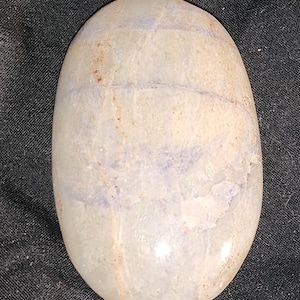 Puede incluir: Una piedra lisa de forma ovalada, de color gris claro con vetas blancas y moradas claras. La piedra tiene un acabado pulido.