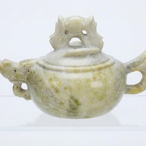 May include: A pale green stone teapot featuring a carved dragon head spout and a matching dragon-shaped handle. The lid is topped with a sculpted creature finial. The teapot has a mottled, semi-translucent appearance with natural stone variations.