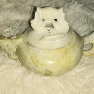 May include: A carved stone teapot with a lid featuring a detailed animal design. The teapot is primarily a pale yellow color with darker yellow and green veining. The handle and spout are curved, and the lid has a decorative animal figure.