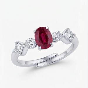 May include: A silver-toned ring featuring a central oval-cut deep red ruby gemstone. The band is accented with two round-cut clear stones and two square-cut clear stones on either side. The ring has an open, adjustable band design at the base.