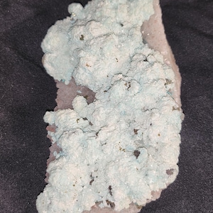May include: A cluster of light blue and white crystals with a rough, textured surface. The crystals are set against a dark background.