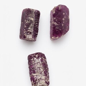 May include: Three raw, unpolished ruby crystals with a deep magenta and purple hue. Each stone features a rough, textured surface with natural crystalline formations and irregular edges, displaying a mix of opaque and translucent deep red tones.