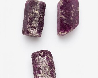 May include: Three raw, unpolished ruby crystals with a deep magenta and purple hue. Each stone features a rough, textured surface with natural crystalline formations and irregular edges, displaying a mix of opaque and translucent deep red tones.