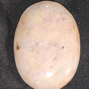 Puede incluir: Una piedra lisa, de forma ovalada y pulida, de color rosa claro y blanco. La piedra tiene un patrón sutil y en espiral y un brillo ligeramente iridiscente.