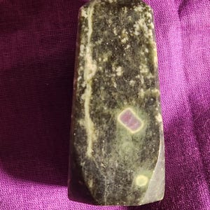 Könnte beinhalten: Eine dunkelgrüne und schwarze Kristallspitze mit einer rosa und weißen Einlagerung. Der Kristall ist poliert und hat eine glatte Oberfläche.