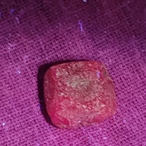 May include: A small, square, red object with a textured surface. The object is set against a purple background. The object appears to be a decorative item or a component of a larger piece.