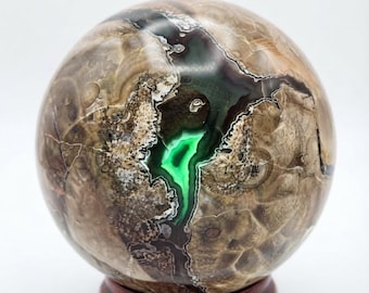 May include: A polished spherical stone featuring a complex pattern of tan, brown, and dark grey swirls. A prominent, jagged fissure runs through the centre, revealing a vibrant, glowing bright green mineral interior with white crystalline edges.