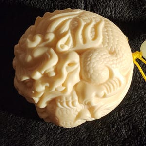 Op de afbeelding: Ivoor-kleurige, gebeeldhouwde hanger met een gedetailleerd drakenontwerp. De hanger is rond en heeft een geel koord. De draak is ingewikkeld gesneden met schubben en een gedetalleerd gezicht. De achtergrond is een donkere, getextureerde stof.