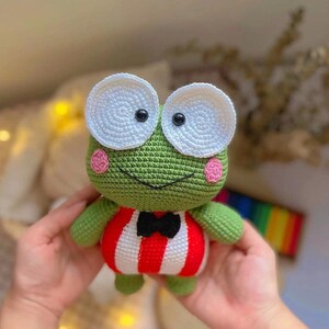 K&ouml;nnte beinhalten: Ein gr&uuml;ner H&auml;kel-Frosch, der ein rot-wei&szlig; gestreiftes Outfit mit einer schwarzen Schleife tr&auml;gt. Der Frosch hat gro&szlig;e wei&szlig;e Augen mit schwarzen Pupillen und rosa Wangen.