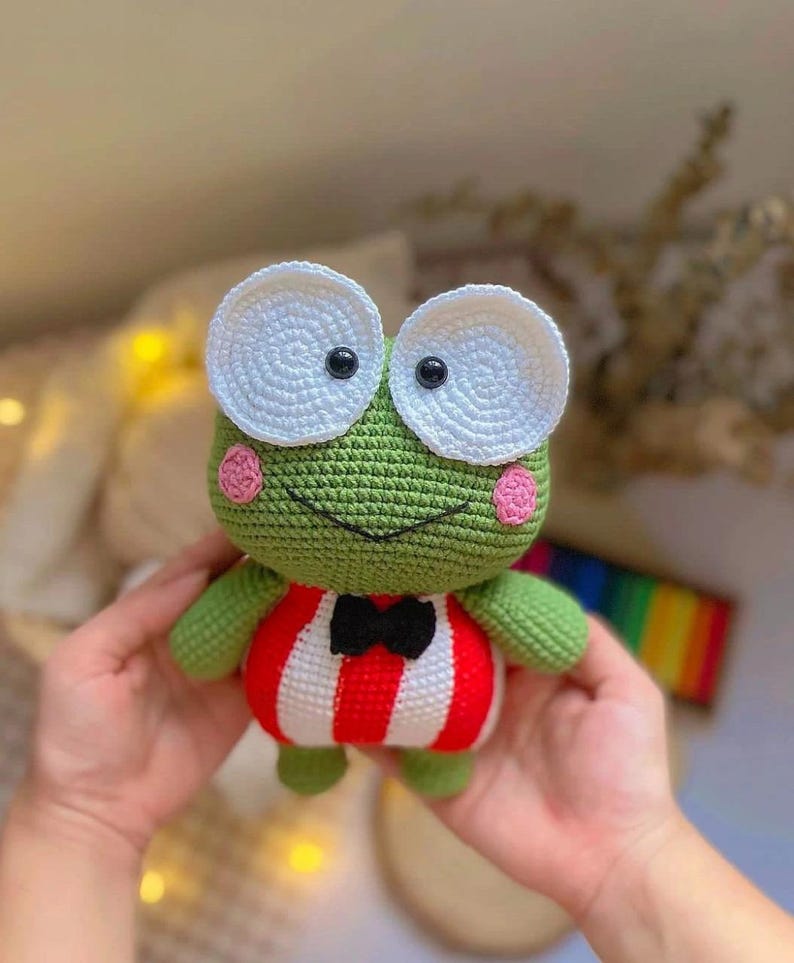 K&ouml;nnte beinhalten: Ein gr&uuml;ner H&auml;kel-Frosch, der ein rot-wei&szlig; gestreiftes Outfit mit einer schwarzen Schleife tr&auml;gt. Der Frosch hat gro&szlig;e wei&szlig;e Augen mit schwarzen Pupillen und rosa Wangen.