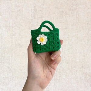 May include: A small green crocheted bag with a white daisy flower on the front. The bag has a handle and is small enough to fit in the palm of a hand.