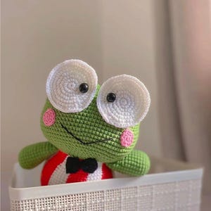 K&ouml;nnte beinhalten: Ein gr&uuml;nes H&auml;kel-Froschspielzeug mit gro&szlig;en wei&szlig;en Augen und rosa Wangen. Der Frosch tr&auml;gt ein rot-wei&szlig; gestreiftes Hemd mit einer schwarzen Fliege. Der Frosch sitzt in einem wei&szlig;en Weidenkorb.