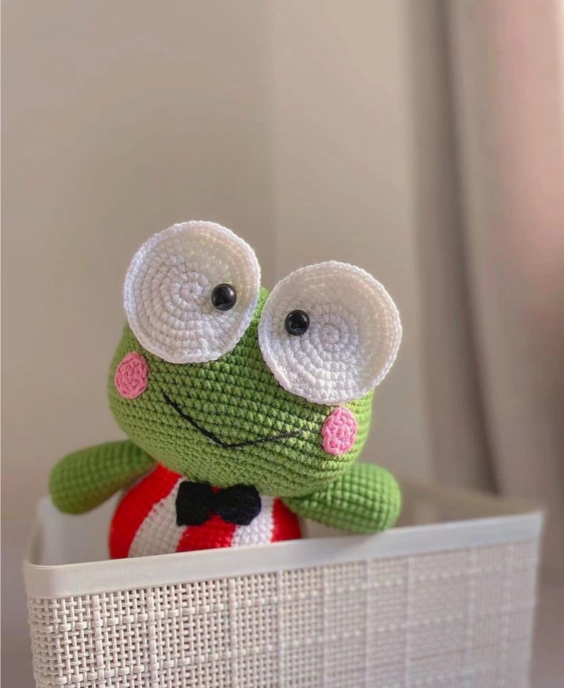 K&ouml;nnte beinhalten: Ein gr&uuml;nes H&auml;kel-Froschspielzeug mit gro&szlig;en wei&szlig;en Augen und rosa Wangen. Der Frosch tr&auml;gt ein rot-wei&szlig; gestreiftes Hemd mit einer schwarzen Fliege. Der Frosch sitzt in einem wei&szlig;en Weidenkorb.