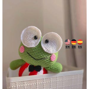 Könnte beinhalten: Ein grüner Häkel-Frosch, der ein rot-weiß gestreiftes Outfit mit einer schwarzen Schleife trägt. Der Frosch hat große weiße Augen mit schwarzen Pupillen und rosa Wangen. Der Frosch sitzt in einem weißen Weidenkorb.