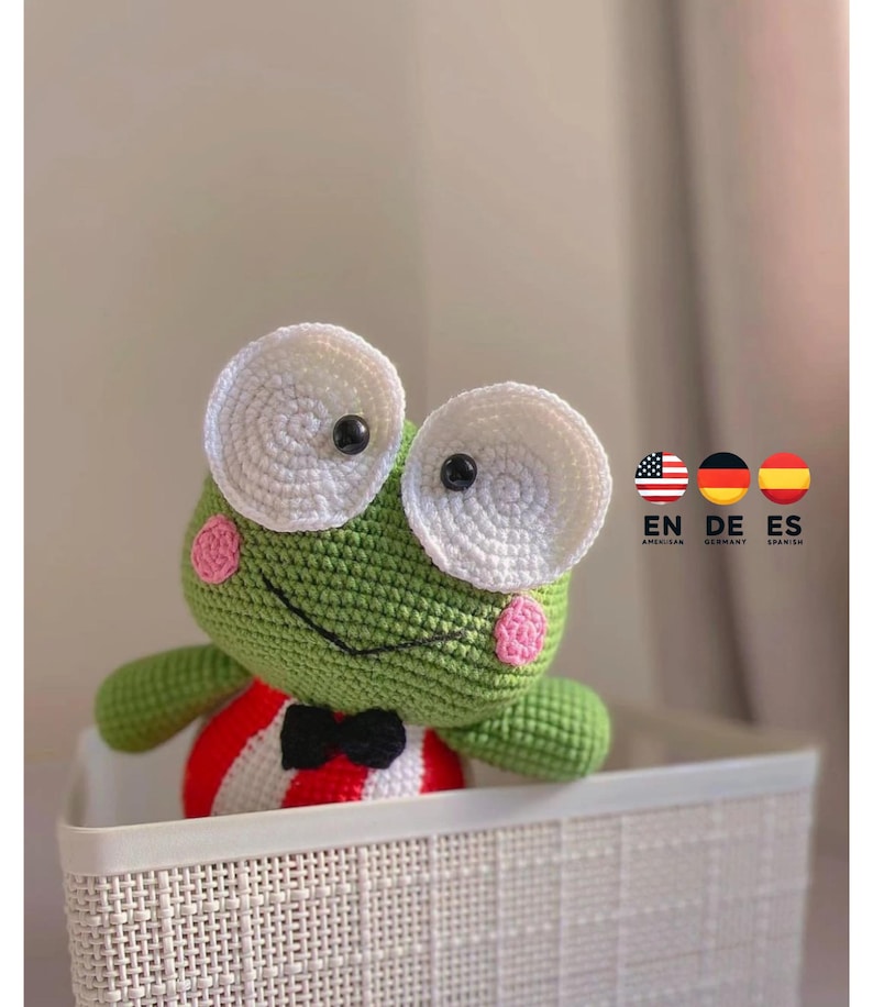 K&ouml;nnte beinhalten: Ein gr&uuml;ner H&auml;kel-Frosch, der ein rot-wei&szlig; gestreiftes Outfit mit einer schwarzen Schleife tr&auml;gt. Der Frosch hat gro&szlig;e wei&szlig;e Augen mit schwarzen Pupillen und rosa Wangen. Der Frosch sitzt in einem wei&szlig;en Weidenkorb.