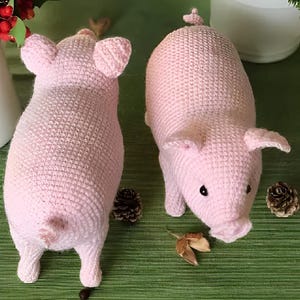 Puede incluir: Dos peluches de cerdo de crochet rosa. Un cerdo está mirando hacia adelante con la cola enroscada. El otro cerdo está mirando hacia atrás con la cola enroscada.
