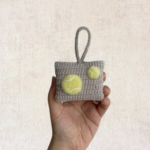 May include: A small gray crocheted bag with a loop handle and two yellow felt tennis balls attached.