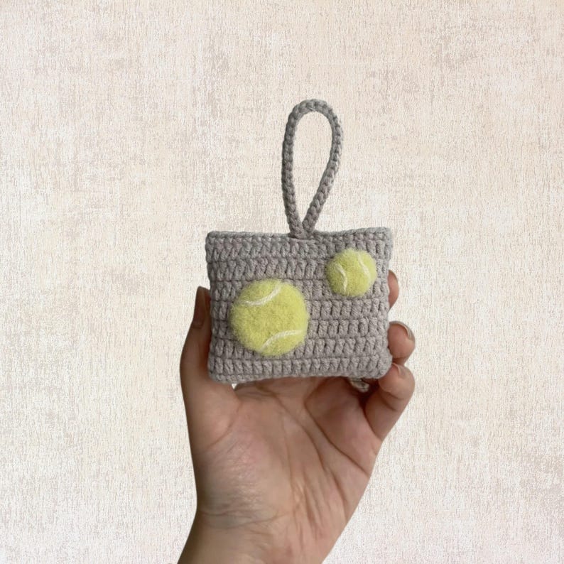 May include: A small gray crocheted bag with a loop handle and two yellow felt tennis balls attached.
