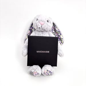 Puede incluir: Un conejo de peluche gris con orejas y pies estampados con flores sostiene una caja negra. La caja tiene la palabra "VIVIDMAISIE" impresa en blanco. El conejo tiene una nariz rosa, sobre un fondo blanco.