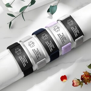 Peut inclure: Plusieurs bracelets d'alerte médicale de couleurs variées, dont noir, gris, marine et lavande. Chaque bracelet comporte une plaque métallique avec un symbole médical et du texte gravé. Le texte comprend un nom, une condition médicale, une date de naissance et "Equipment Inside."