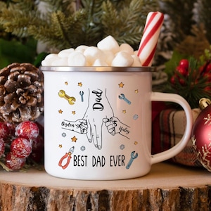 Op de afbeelding: Witte emaille mok met een handvat, gevuld met marshmallows en een zuurstok. De mok heeft een afbeelding van handen die elkaar vasthouden, het woord "Dad" en de tekst "BEST DAD EVER". Het heeft ook de namen "Ozden" en "Haley" en gereedschapsillustraties.