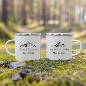 Puede incluir: Dos tazas de esmalte blanco con bordes plateados, con un diseño de cordillera negra y el texto "Aubrey & Nicky May 12,2025". Las tazas están sobre hierba y musgo verdes, con un fondo borroso de bosque.