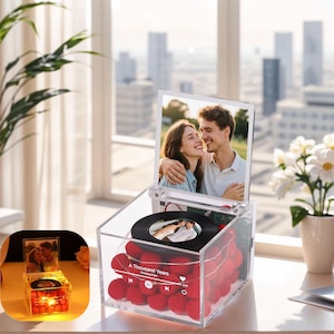 Pode incluir: Caixa de acrílico transparente com uma foto de um casal, um pequeno disco com uma foto e pompons vermelhos. A caixa tem uma foto do casal e o texto "A Thousand Years".