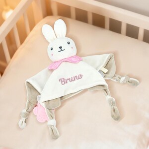 Puede incluir: Una manta de seguridad para bebé blanca y gris con una cabeza de conejo y el nombre "Bruno" bordado en rosa. El conejo tiene mejillas rosas y un cuello rosa. La manta está sobre una superficie rosa.